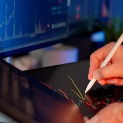 Person using a stylus on a tablet for stock market analysis, with charts on a monitor.